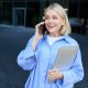 Portrait of young office manager, woman answers phone call, holding laptop, standing near office building, talking to someone over smartphone.